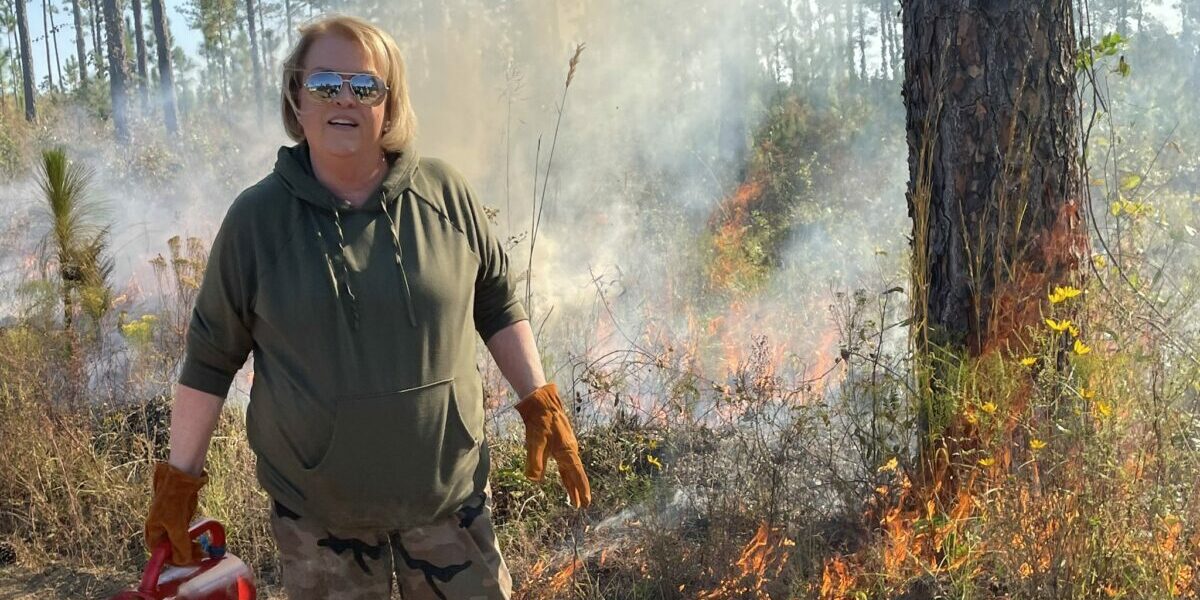 A woman stands with a prescribed fire burning behind her.
